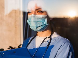 Young sad female caucasian UK US GP EMS doctor carer looking through ICU window,fear uncertainty in eyes,wearing face mask gazing at sun,hope faith in overcoming Coronavirus COVID-19 pandemic crisis