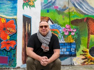 Rob Linn茅 sits on the curb in front of a colorful mural depicting life in El Salvador red hibiscus flowers with a frog one of them, a woman pupusa (the national dish of El Salvador) over an open fire, a llama, a mountain, and a garden wall with a vase of pink tulips on its top.