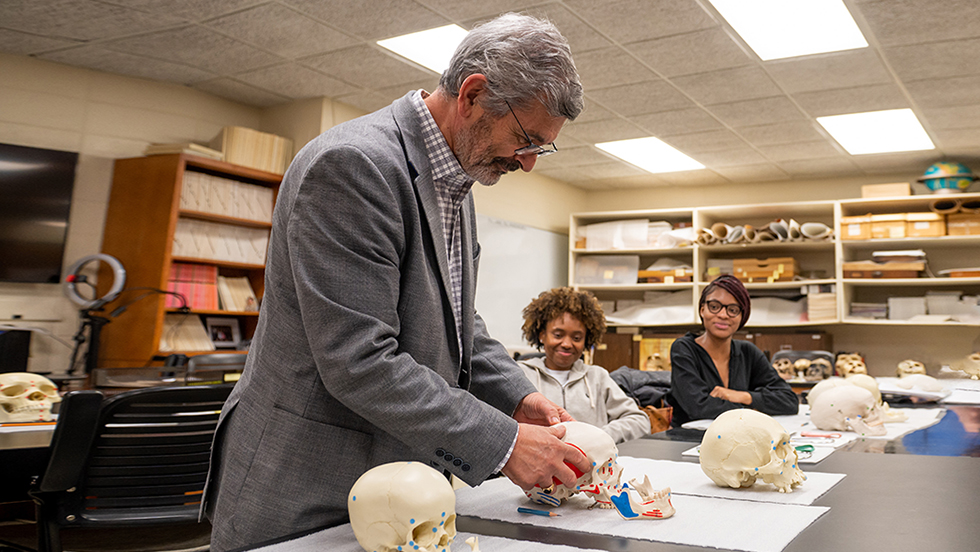 A photograph of Professor Anagnostis Agelarakis, PhD, examining one of three skulls on a countertop in his lab. Two of his students look on.