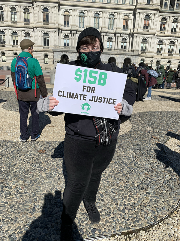 Kelly Andreuzzi, wearing a Covid mask, holds a sign saying "$15 billion for climate justice." A "NY Renews" logo is on the bottom of the sign. Kelly is in front of the New York capitol building in Albany. Others at the rally are standing behind her.
