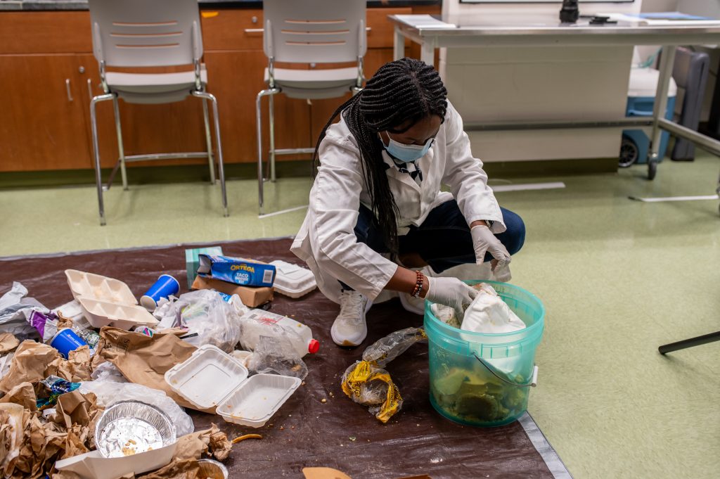 Morenike Olushola-Oni in her lab, separating food waste. She is on the floor, and putting food waste into a bucket while other trash鈥攊ncluding styrofoam clamshells trays, aluminum tins, plastic cups and bags used to store food鈥攊s heaped on a tarp.