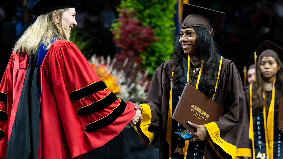 A student receives her diploma and a congratulatory handshake from a faculty member at Adelphi's graduation ceremony. Two students are behind her, waiting for their turn to receive their diploma.