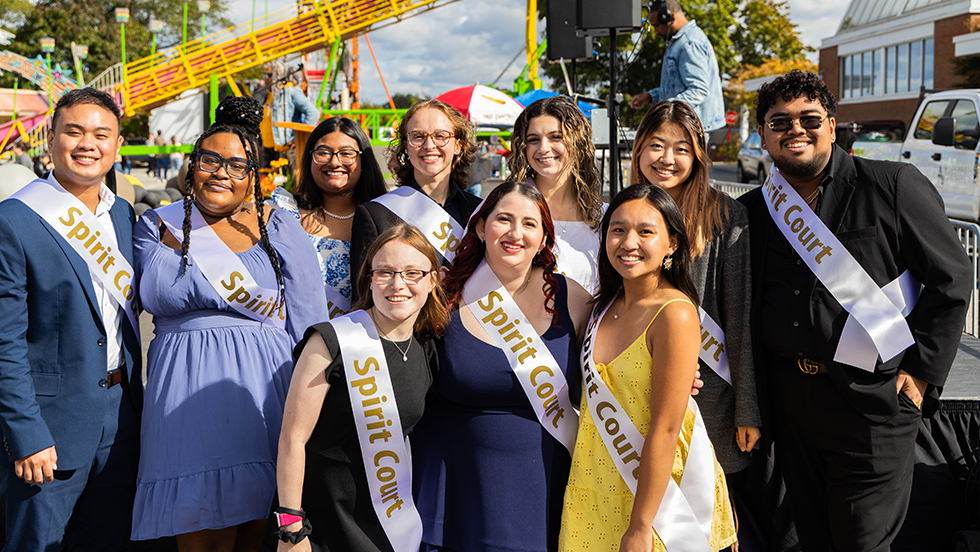 10 students of different ethnicities are smiling, standing against a carnival backdrop, and wear sashes that read "Spirit Court"