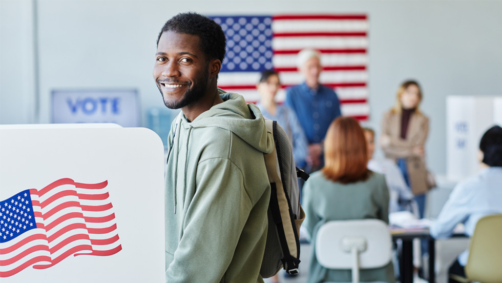 A person standing in front of a voting table with an American flag behind him