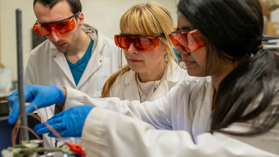 Professor Widera-Kalinowska and two students working in a campus lab. All are wearing white lab coats and orange protective glasses. One of the students is wearing blue exam gloves.