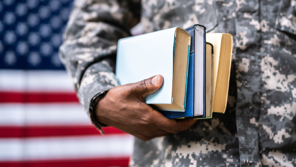  Close-up of a Black man's hand holding textbooks against his camouflage clothing with American flag in background
