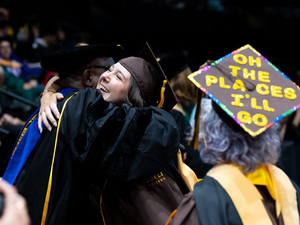 A happy graduate hugs her professor on Commencement day. An older graduate wears a cap that says "Oh, the Places I'll Go."