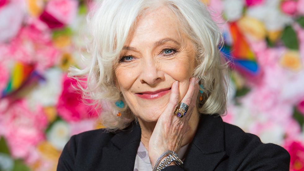A woman with white hair and blue eyes is slightly smiling, standing against a blurred backdrop of flowers 